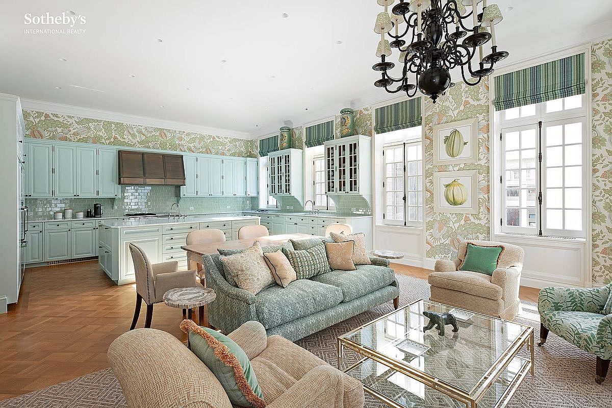 This elegant, open-concept kitchen and living area features a sophisticated blend of light blue cabinetry, patterned wallpaper, and warm hardwood flooring. A large central island serves as a focal point, complemented by a comfortable seating area with a glass-topped coffee table and a striking black chandelier overhead. The space is bathed in natural light from tall, classic windows, creating a bright, airy, and luxurious atmosphere perfect for both cooking and entertaining.