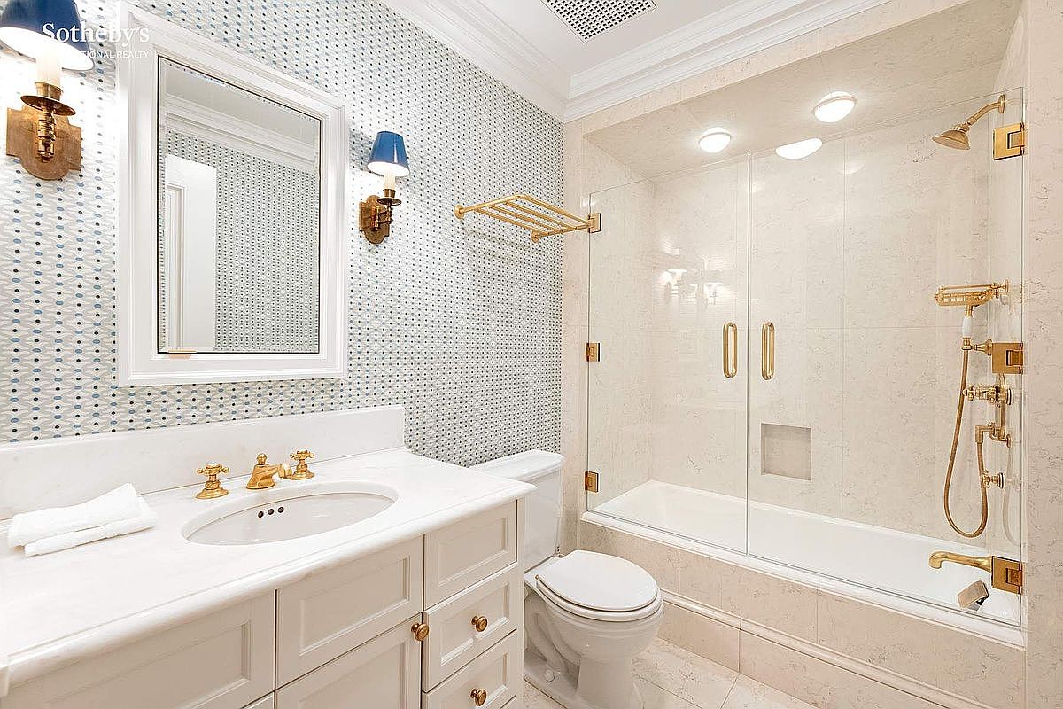This elegant bathroom features a classic vanity with a white countertop, gold-toned fixtures, and a distinctive patterned wallpaper that adds a touch of sophistication. A glass-enclosed shower-tub combination with matching gold hardware and recessed lighting creates a bright, clean, and luxurious atmosphere. The perspective is a wide-angle shot capturing the entire functional layout, emphasizing the high-end finishes and polished aesthetic.