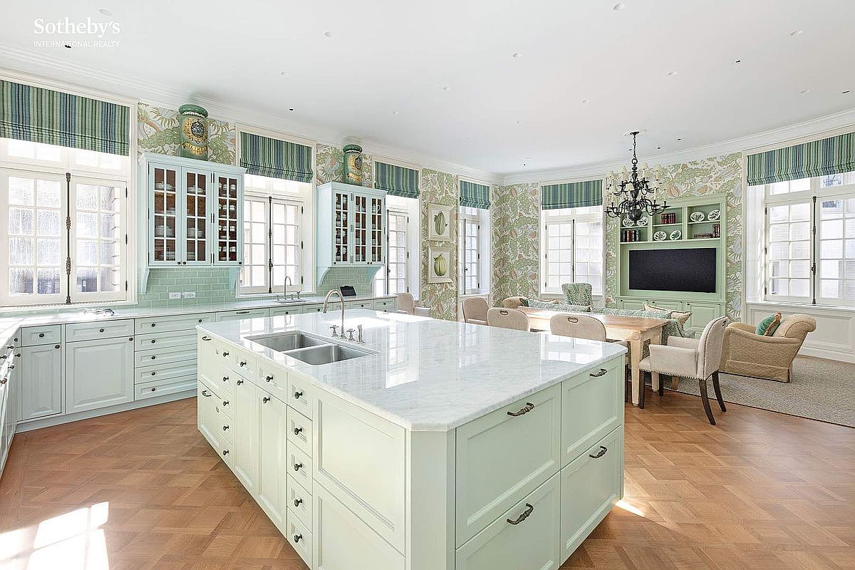 This elegant, light-filled kitchen features a large central island with a white marble countertop and pale green cabinetry, creating a sophisticated and airy atmosphere. The space seamlessly integrates a dining area with patterned wallpaper, classic chandeliers, and large windows that flood the room with natural light. The herringbone wood flooring and refined architectural details contribute to a luxurious, timeless aesthetic perfect for high-end living.
