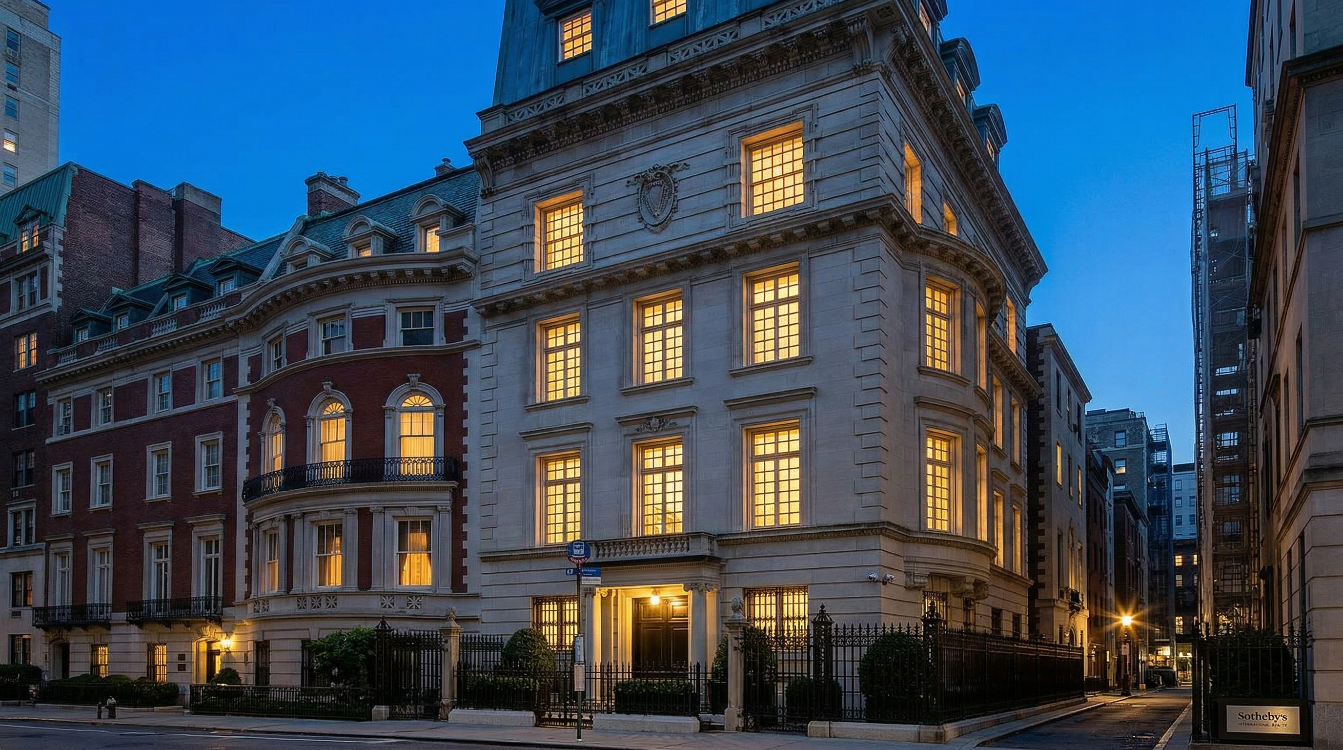 This image captures the grand, symmetrical front facade of a classic limestone townhouse, showcasing elegant Beaux-Arts architectural details. The structure features a prominent mansard roof with dormer windows, a decorative central crest, and a stately entrance framed by a wrought-iron fence. The perspective is a direct, eye-level shot that emphasizes the building's imposing scale and sophisticated urban presence.