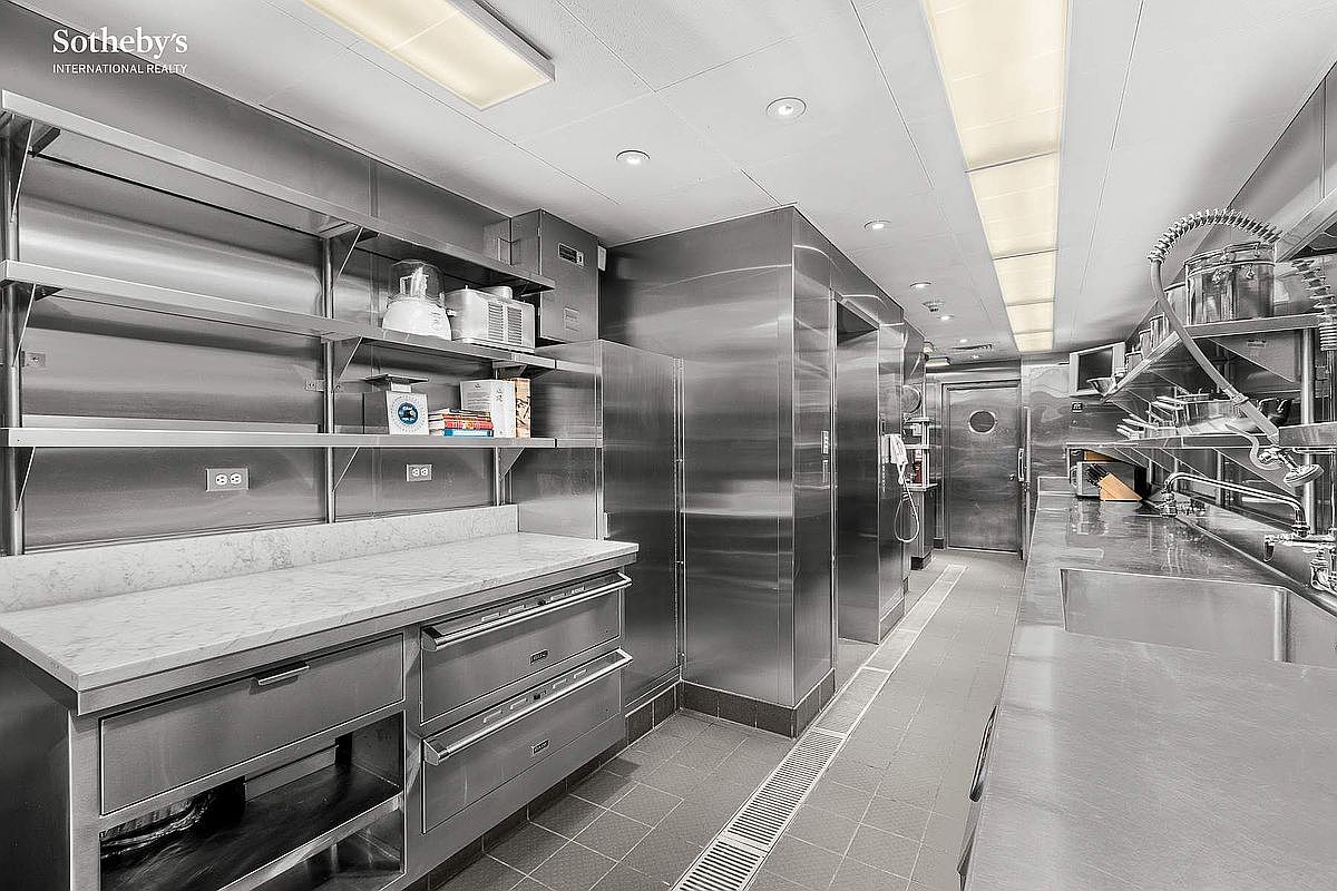 This professional-grade commercial kitchen features extensive stainless steel surfaces, industrial-style shelving, and high-end appliances, creating a sleek and functional culinary workspace. The long, narrow layout is illuminated by recessed lighting and overhead panels, emphasizing the clean, utilitarian aesthetic. The perspective captures the full depth of the room, highlighting the seamless integration of heavy-duty equipment and ample counter space.