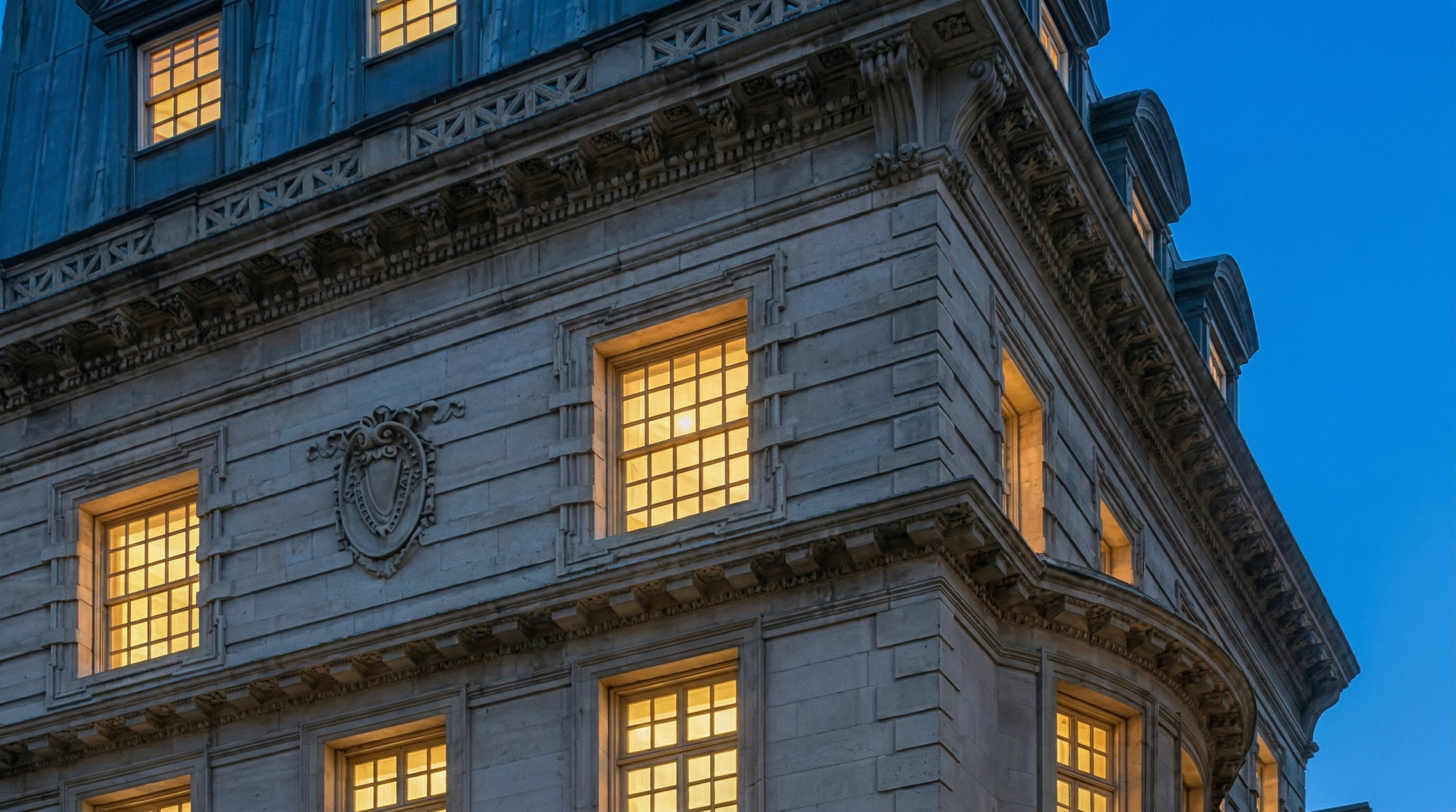 This image shows the upper levels of a grand, classical stone building during the dusk hours. The architectural features, including ornate stonework, sash windows with a warm interior glow, and a slate-tiled mansard roof with dormers, convey a sense of historic luxury and sophistication.