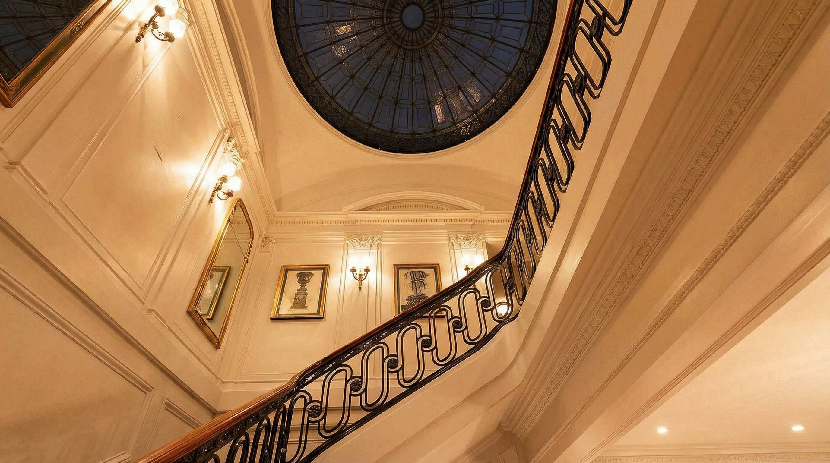 This grand foyer features an elegant, sweeping staircase with an ornate black metal railing and a wood handrail. Above, a circular stained-glass dome creates a dramatic architectural centerpiece, providing soft, ambient light throughout the space. The area is decorated with classic framed art and wall sconces, evoking a sophisticated, traditional aesthetic.