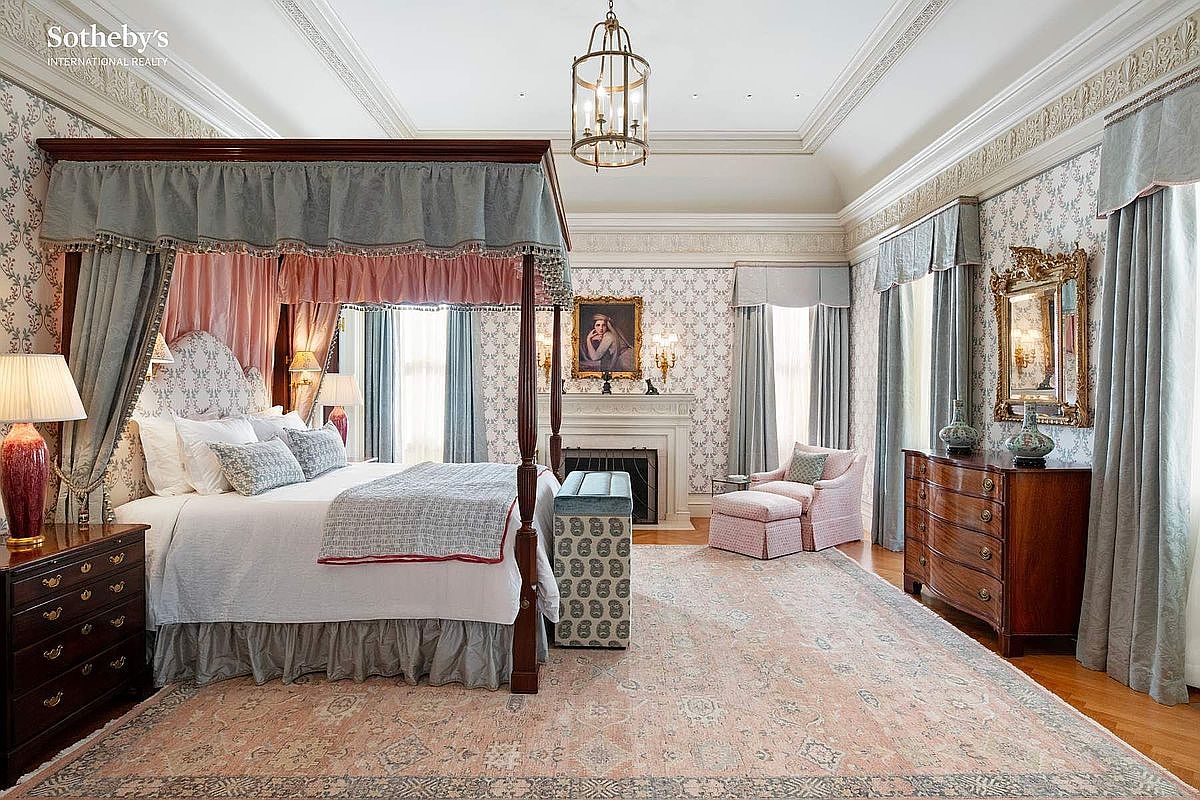 This elegant primary bedroom features a grand four-poster canopy bed as the centerpiece, complemented by sophisticated floral wallpaper and ornate crown molding. The room includes a cozy sitting area with a patterned armchair, a fireplace with a framed portrait above, and a rich wooden dresser, all tied together by a large, soft-toned area rug. The perspective is a wide, eye-level shot that captures the luxurious, traditional aesthetic and spacious layout of the suite.