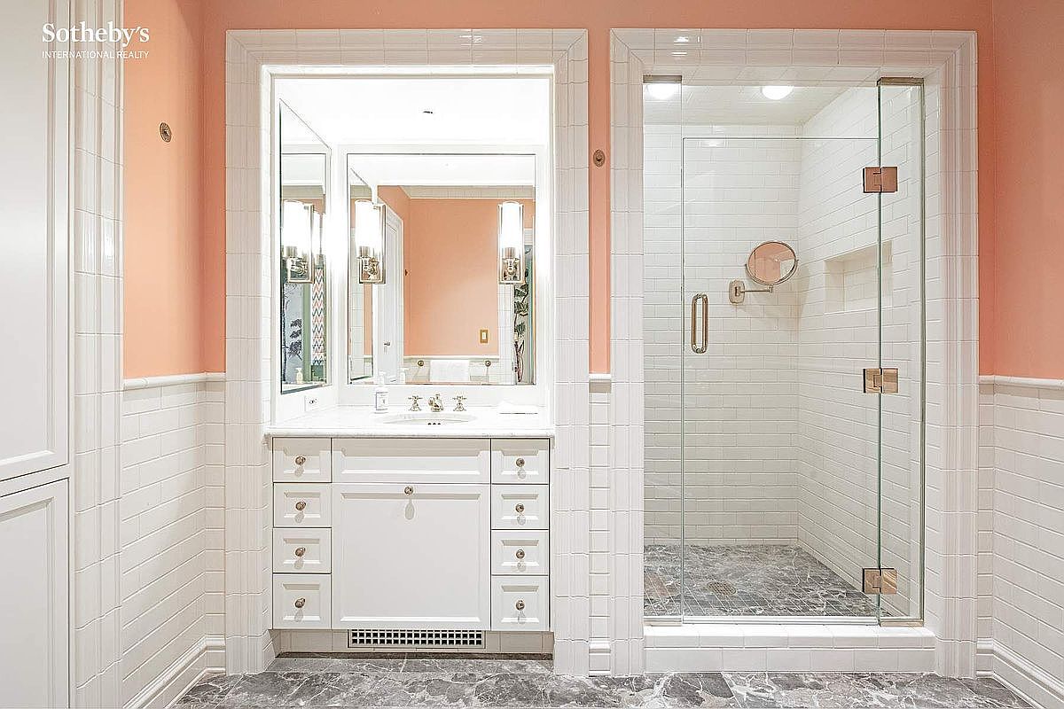 This elegant bathroom features a classic vanity with white cabinetry and a marble countertop, centered beneath a large, framed mirror. To the right, a spacious walk-in shower is enclosed by glass doors and finished with white subway tile walls and a dark marble floor. The room is accented by soft peach-colored walls and white wainscoting, creating a bright, sophisticated, and timeless aesthetic.