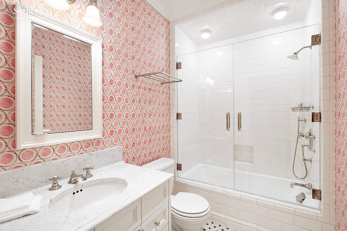 This charming bathroom features a distinctive pink and white circular patterned wallpaper that creates a playful, vintage-inspired aesthetic. The space includes a white marble-topped vanity, a classic white toilet, and a glass-enclosed shower-tub combination with white subway tile walls. The perspective is a wide-angle shot capturing the entire functional layout, highlighting the clean, bright, and well-maintained condition of the room.
