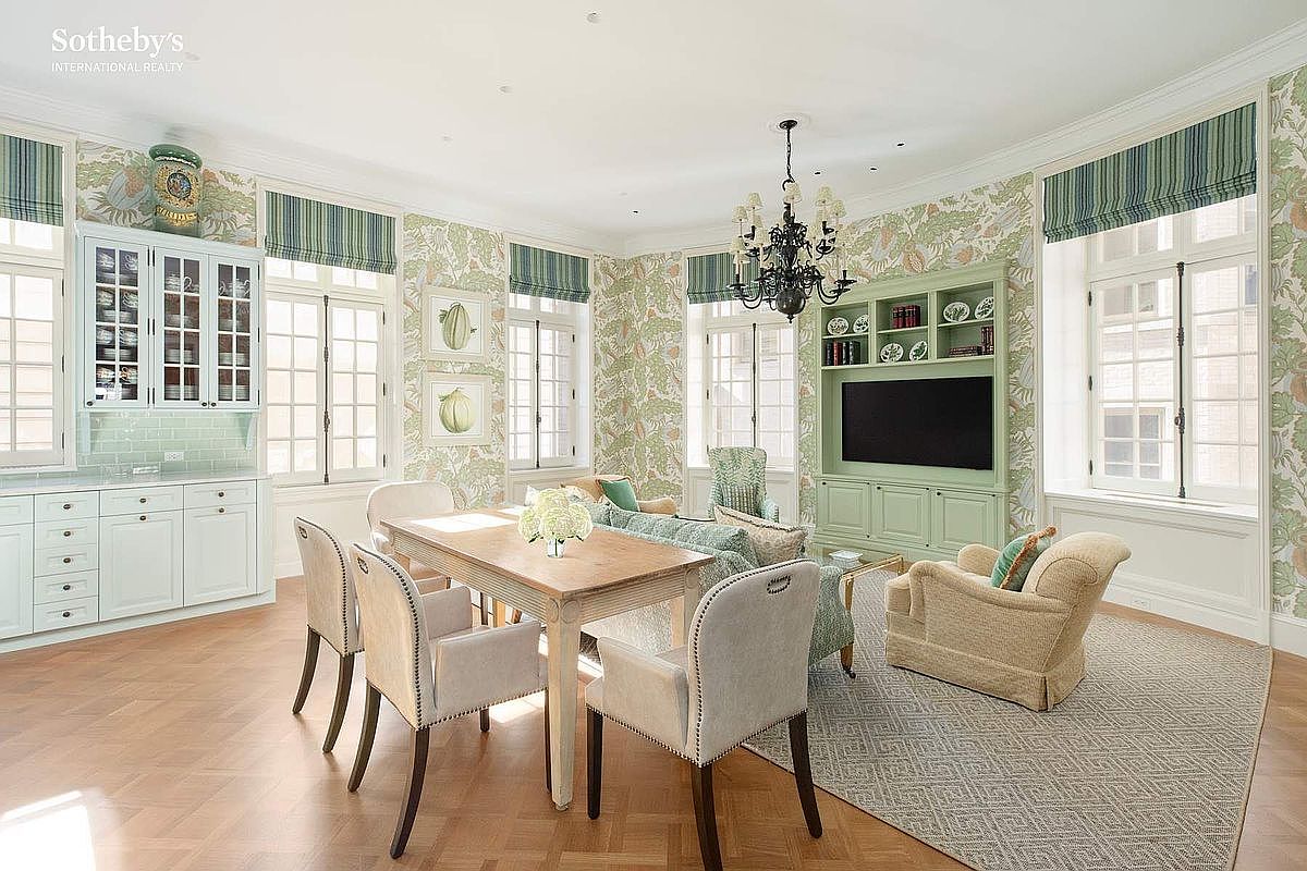 This elegant living room features a sophisticated blend of traditional and transitional design, highlighted by intricate botanical wallpaper and light green built-in cabinetry. The space is anchored by a wooden dining table and a comfortable seating area with plush armchairs, all set upon a textured area rug over herringbone hardwood floors. Large windows with striped Roman shades flood the room with natural light, creating a bright, airy, and refined atmosphere.