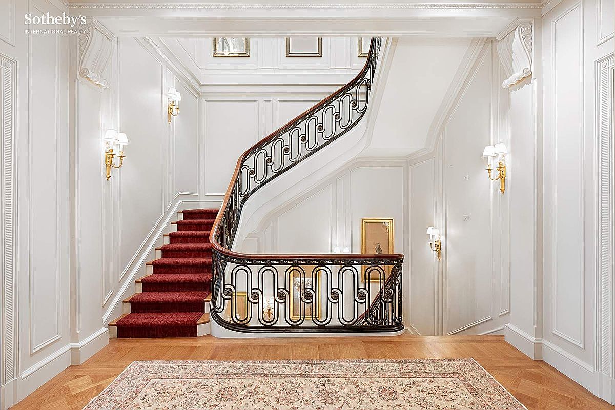 This elegant grand foyer features a sweeping staircase with a rich red runner and an ornate, dark metal railing. The space is characterized by classic architectural details, including crisp white wainscoting, wall sconces, and a polished hardwood floor adorned with a traditional area rug. The perspective is centered, capturing the sophisticated and formal atmosphere of a luxury residence.