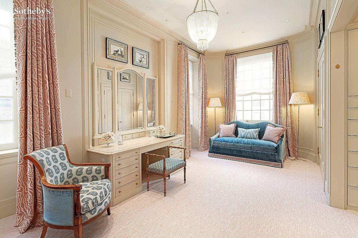 This elegant dressing room or primary bedroom suite features a sophisticated vanity area with a large tri-fold mirror and a plush blue velvet settee positioned by the window. The room is decorated in a refined, traditional style with soft pink patterned curtains, intricate wall molding, and a crystal chandelier that adds a touch of luxury. The perspective is a wide-angle shot that captures the harmonious balance between the functional vanity space and the comfortable seating area.