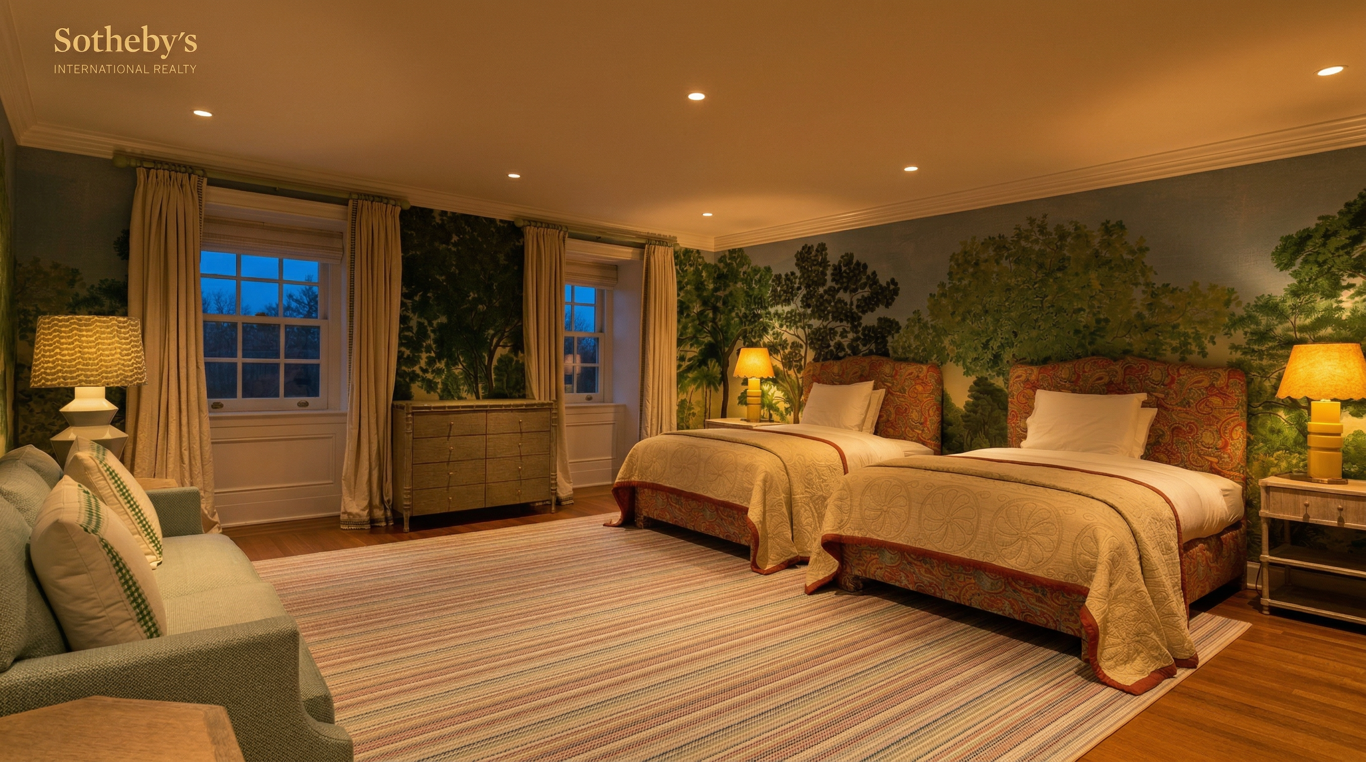 This spacious guest bedroom features two twin beds with ornate paisley-patterned headboards and matching bed skirts, set against a striking floor-to-ceiling mural of a lush forest landscape. The room is brightened by large windows draped in neutral curtains, a light-colored area rug, and a comfortable sofa in the foreground, creating a serene and sophisticated atmosphere. The perspective is a wide-angle shot from the corner of the room, capturing the full layout and the elegant, classic design aesthetic.