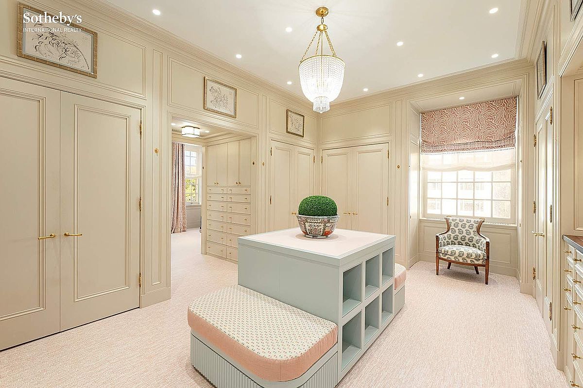 This elegant walk-in closet features custom floor-to-ceiling cabinetry with a soft, neutral finish and a central island providing both storage and seating. A crystal chandelier serves as a sophisticated focal point, while a patterned armchair by the window adds a touch of classic charm. The space is bright and airy, conveying a sense of luxury and organized refinement.