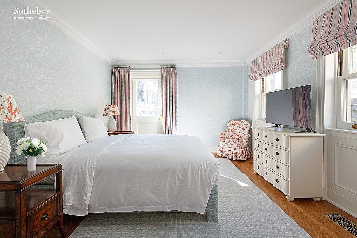 This bright and airy primary bedroom features a classic aesthetic with light blue patterned wallpaper, a plush white-covered bed, and a matching white dresser topped with a flat-screen television. A cozy floral-patterned armchair sits in the corner near windows dressed in striped Roman shades, while a wooden nightstand with a lamp adds a touch of warmth. The room is grounded by a soft grey area rug over hardwood floors, creating a serene and inviting atmosphere.