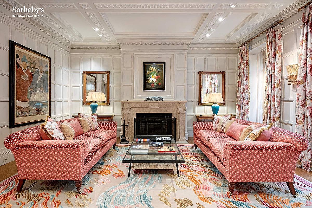 This elegant, formal living room features symmetrical design with two matching coral-patterned sofas facing each other across a glass-topped coffee table. The space is anchored by a grand stone fireplace, flanked by matching mirrors and lamps, all set against sophisticated white wood-paneled walls. A vibrant, abstract-patterned area rug adds a modern touch to the traditional architectural details, creating a refined and inviting atmosphere.