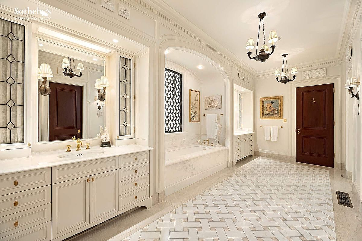 This luxurious primary bathroom features a sophisticated, classic design with a double vanity, marble countertops, and a deep soaking tub nestled in an arched alcove. The space is adorned with elegant chandeliers, intricate tile flooring, and dark wood doors, creating an atmosphere of refined opulence. The wide-angle perspective captures the symmetry and high-end finishes that define this grand, spa-like retreat.