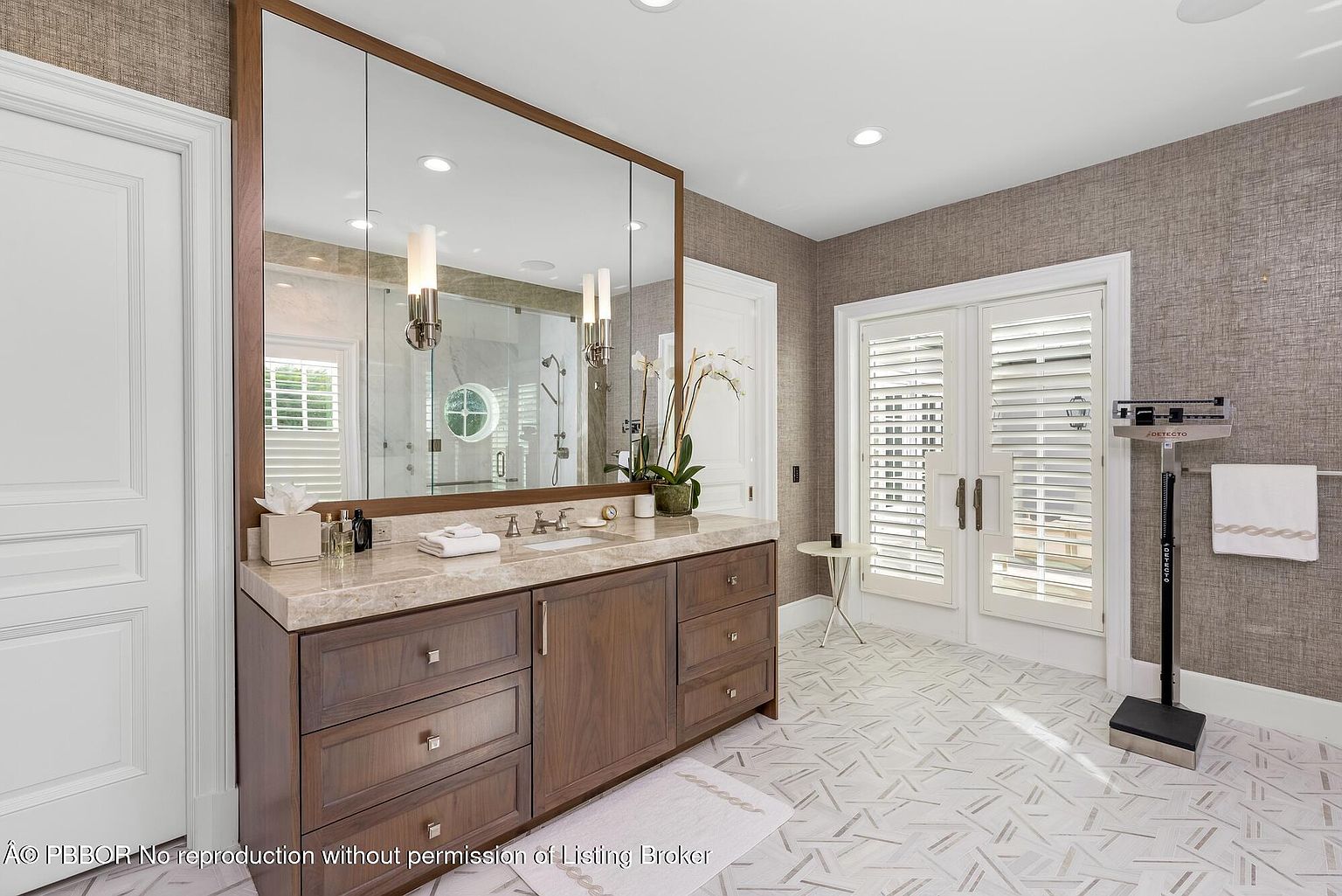 This is a well-lit primary bathroom featuring a large vanity with a marble countertop and dark wood cabinetry. A large mirror reflects a glass-enclosed shower. The room is decorated in neutral tones with textured wallpaper and geometric tile flooring, creating a luxurious and serene atmosphere.