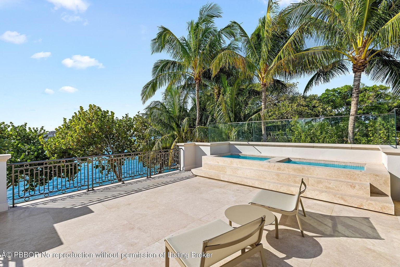 This image showcases a luxurious outdoor patio or balcony area with a built-in spa or plunge pool. The space features elegant stone flooring, modern outdoor furniture, and a decorative wrought iron railing overlooking a serene body of water. Lush palm trees and greenery provide a tropical backdrop, enhancing the property's appeal.