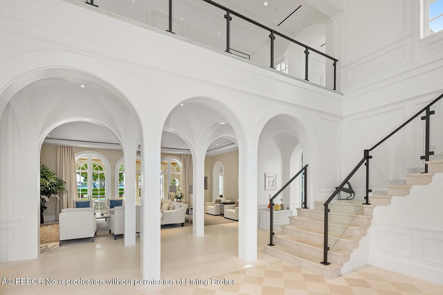 This is an interior shot of a grand hallway featuring a staircase with glass railings and black metal accents. The hallway is characterized by its arched doorways, leading into adjacent living spaces, and a light-colored tile floor. The overall impression is one of spaciousness and elegant design, creating a welcoming and luxurious atmosphere.