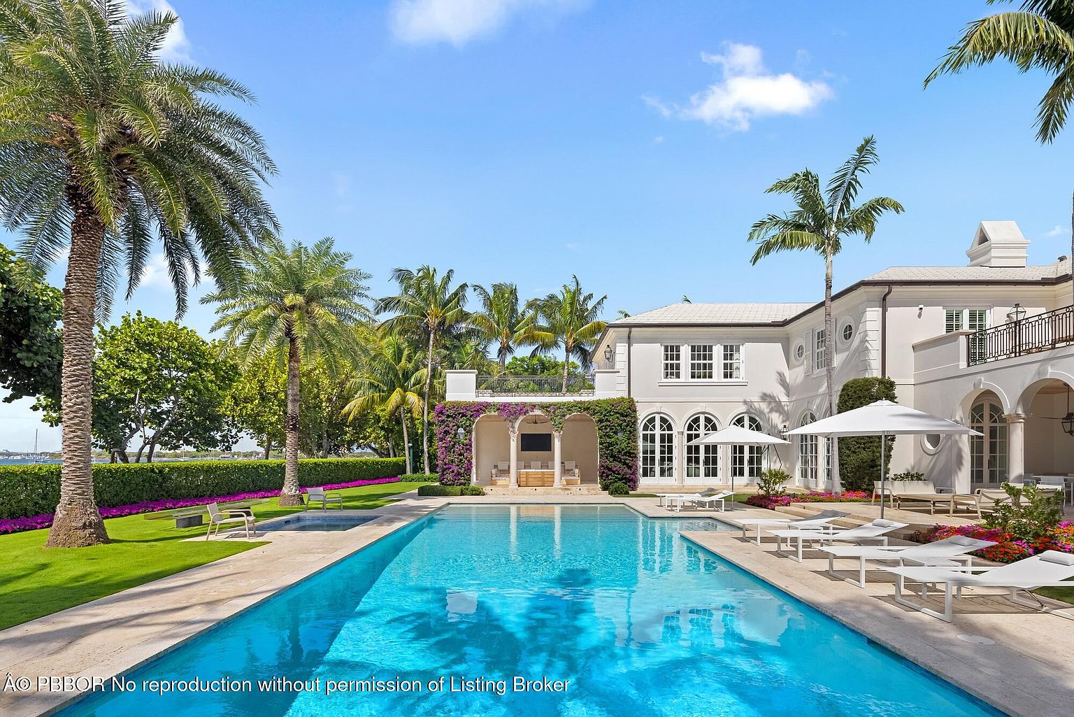 This image showcases a luxurious backyard featuring a large, rectangular swimming pool with crystal-clear blue water. The pool is surrounded by a spacious patio with multiple lounge chairs and umbrellas, perfect for relaxation. The backdrop includes a stunning white mansion with arched windows and lush tropical landscaping, creating an opulent and serene atmosphere.