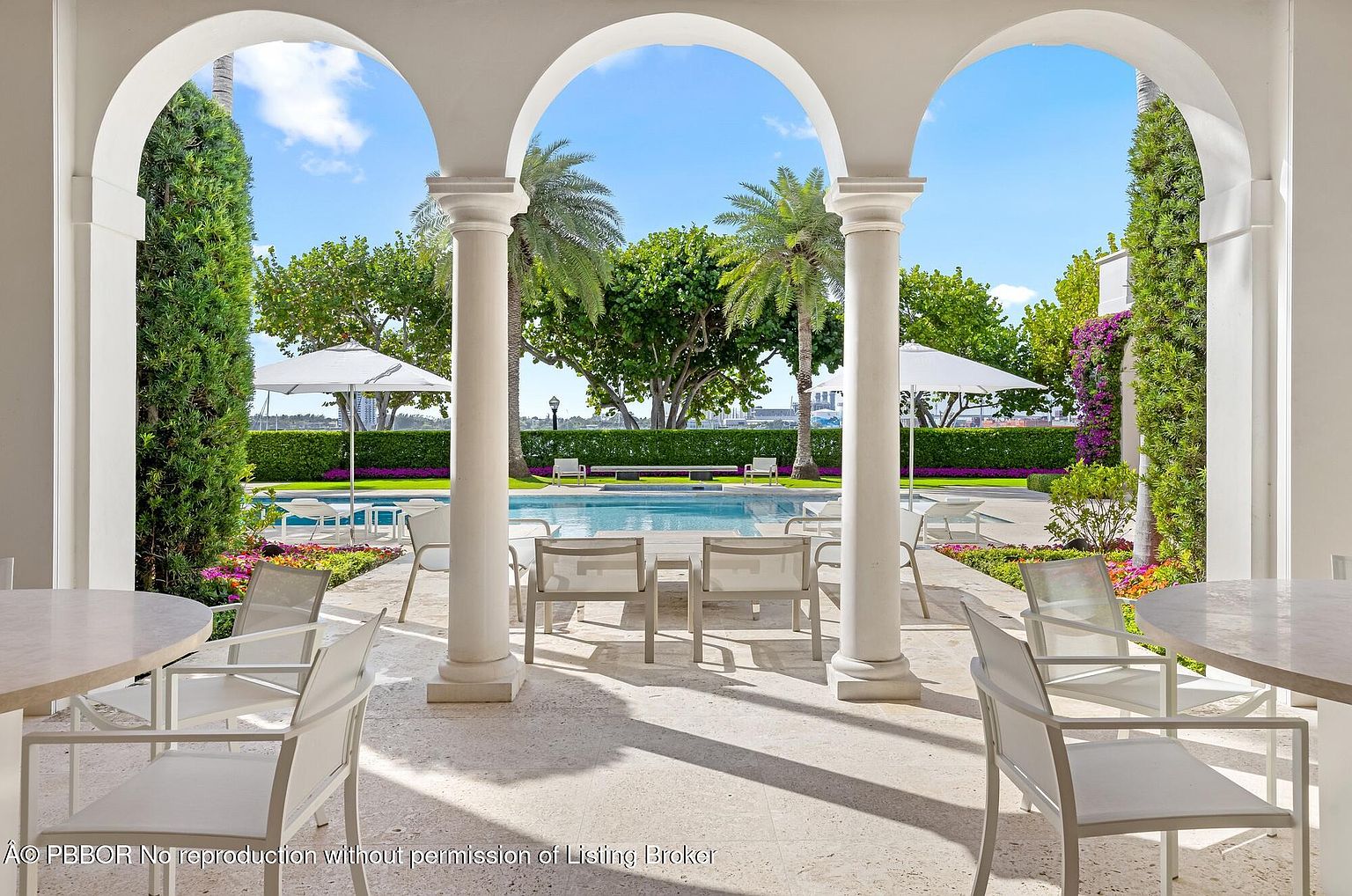 This image showcases a luxurious outdoor pool area framed by elegant white arches and columns. The pool is surrounded by lounge chairs and tables, creating a relaxing and inviting atmosphere. Lush greenery and palm trees add to the tropical ambiance, suggesting a high-end property perfect for entertaining and leisure.