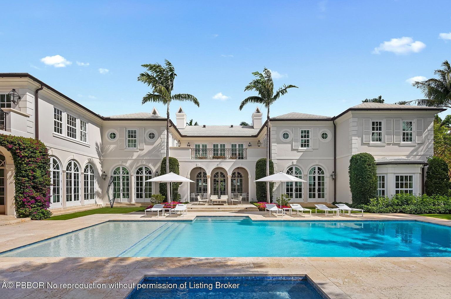 This image showcases the rear exterior of a luxurious home, emphasizing the pool and outdoor living space. The architecture features a symmetrical design with arched windows and doors, complemented by palm trees and manicured landscaping. The pool's clear blue water invites relaxation, while the surrounding lounge chairs suggest an ideal setting for outdoor entertainment.