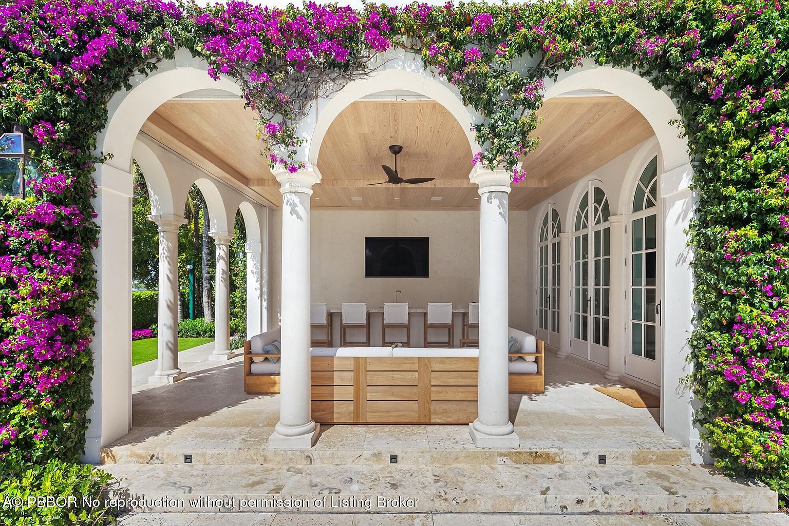 This image showcases a luxurious outdoor patio area, featuring elegant white columns and arches adorned with vibrant purple bougainvillea. The space includes comfortable seating arrangements, a large flat-screen television, and a ceiling fan, creating an inviting atmosphere for relaxation and entertainment. The design seamlessly blends indoor and outdoor living, highlighting the property's upscale appeal.