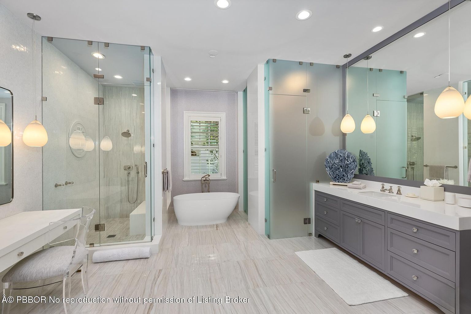 This is a luxurious primary bathroom featuring a double vanity with gray cabinetry and white countertops, illuminated by pendant lighting. A freestanding bathtub sits beneath a window with shutters, and there are two separate glass-enclosed showers. The flooring is light-colored tile, contributing to a bright and spacious feel.