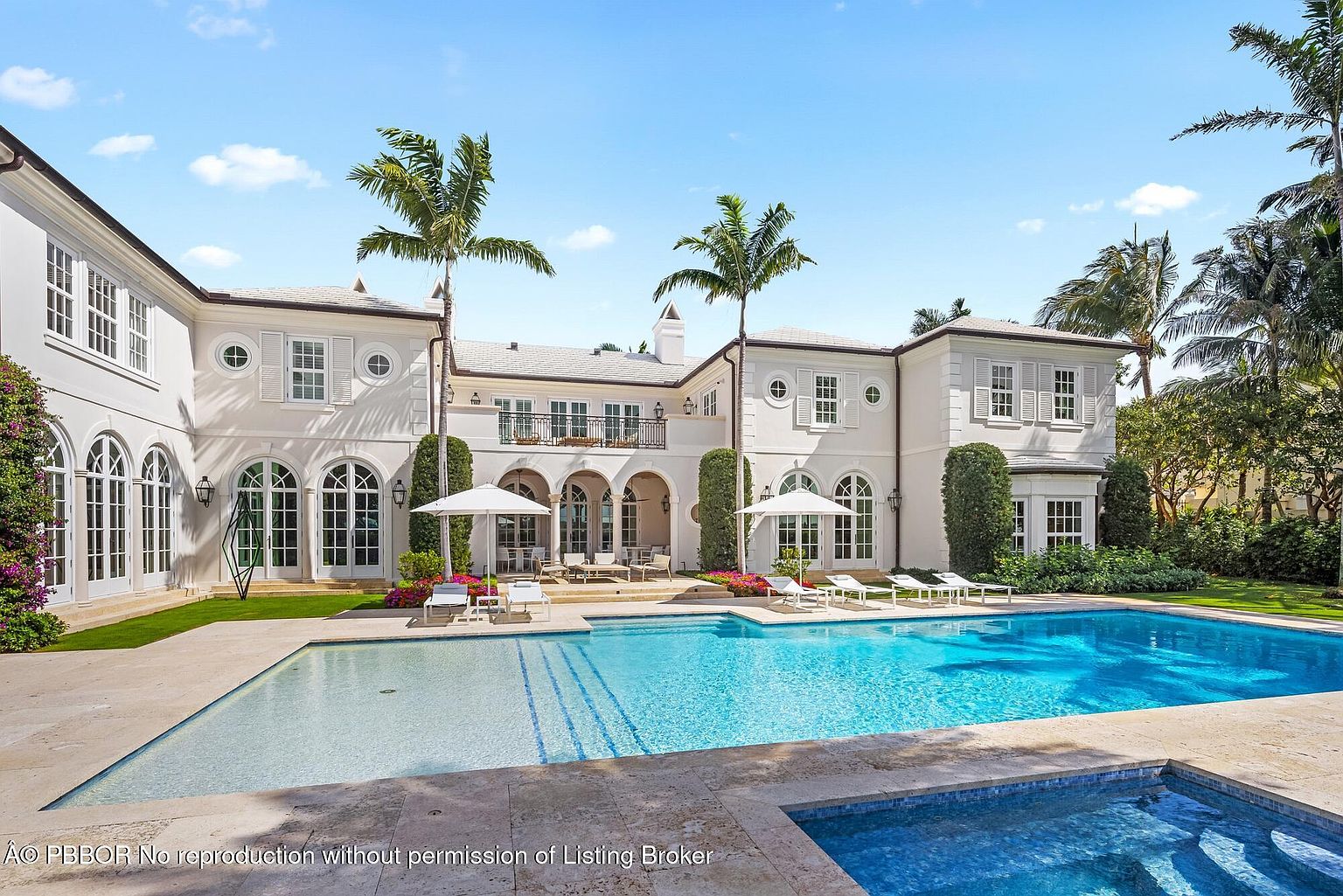 This image showcases the rear exterior of a luxurious home, emphasizing the pool and spa area. The large, rectangular pool features clear blue water and is surrounded by a stone patio with lounge chairs and umbrellas. The house itself is a grand, white structure with arched windows, a balcony, and meticulously maintained landscaping, creating an inviting and opulent outdoor living space.
