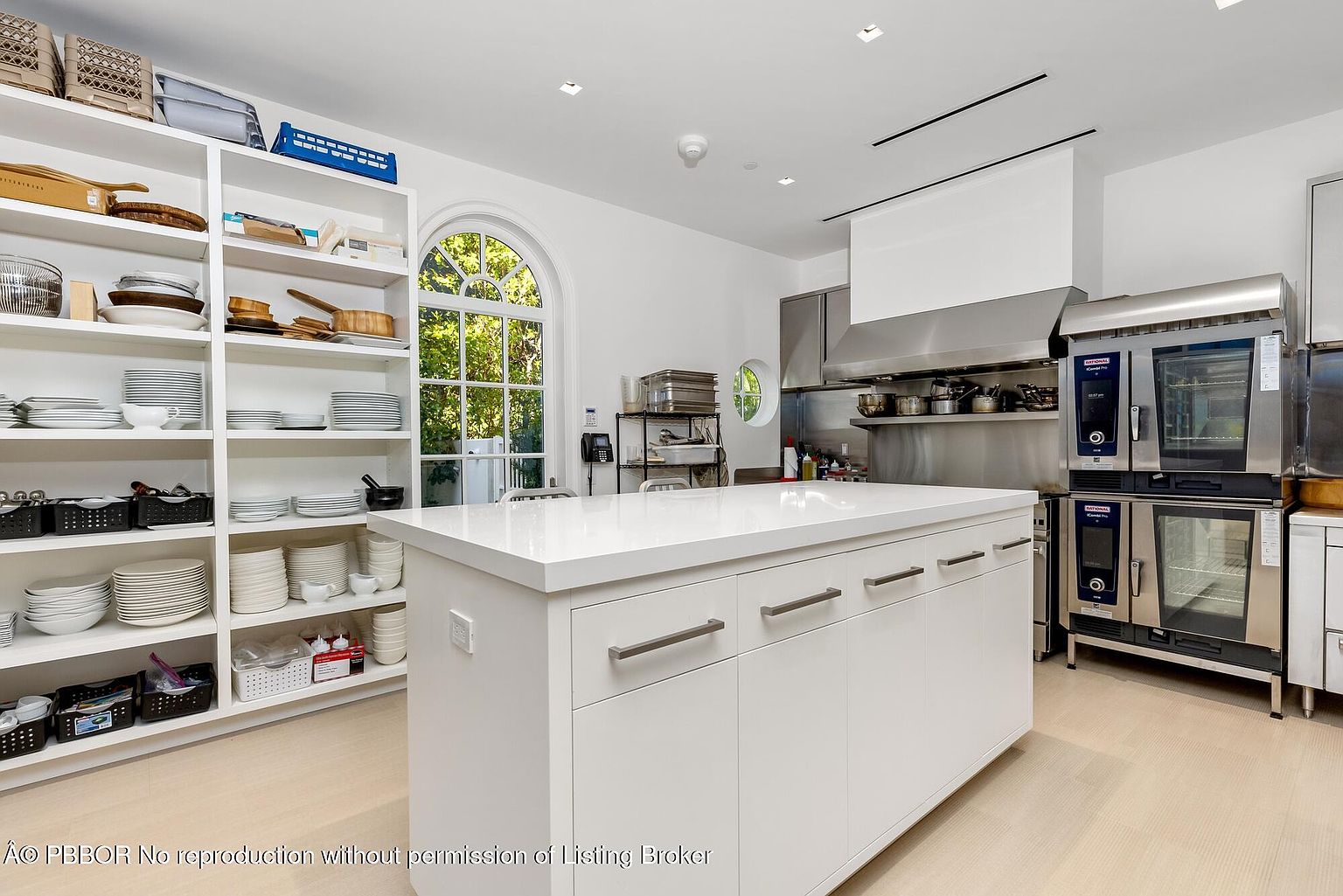 This is a bright and spacious kitchen featuring a large white island with modern hardware, providing ample counter space and storage. Stainless steel appliances, including professional-grade ovens and a ventilation system, suggest a high-end culinary setup. A large arched window and open shelving filled with dishware add to the kitchen's appeal, creating a functional and aesthetically pleasing space.