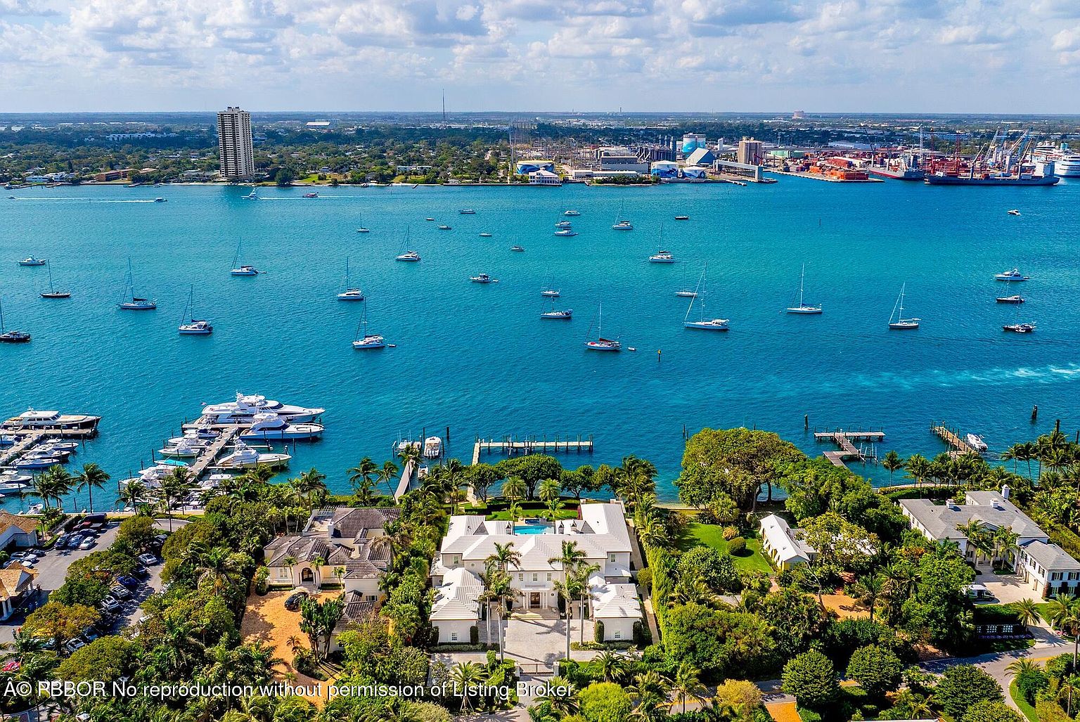 This aerial view showcases a luxurious waterfront property with a grand white mansion featuring a symmetrical design, a central courtyard with a pool, and lush landscaping. The property includes a private dock with yachts and boats, set against a backdrop of a vibrant blue bay dotted with sailboats and a distant cityscape. The overall impression is one of opulence and serene coastal living.