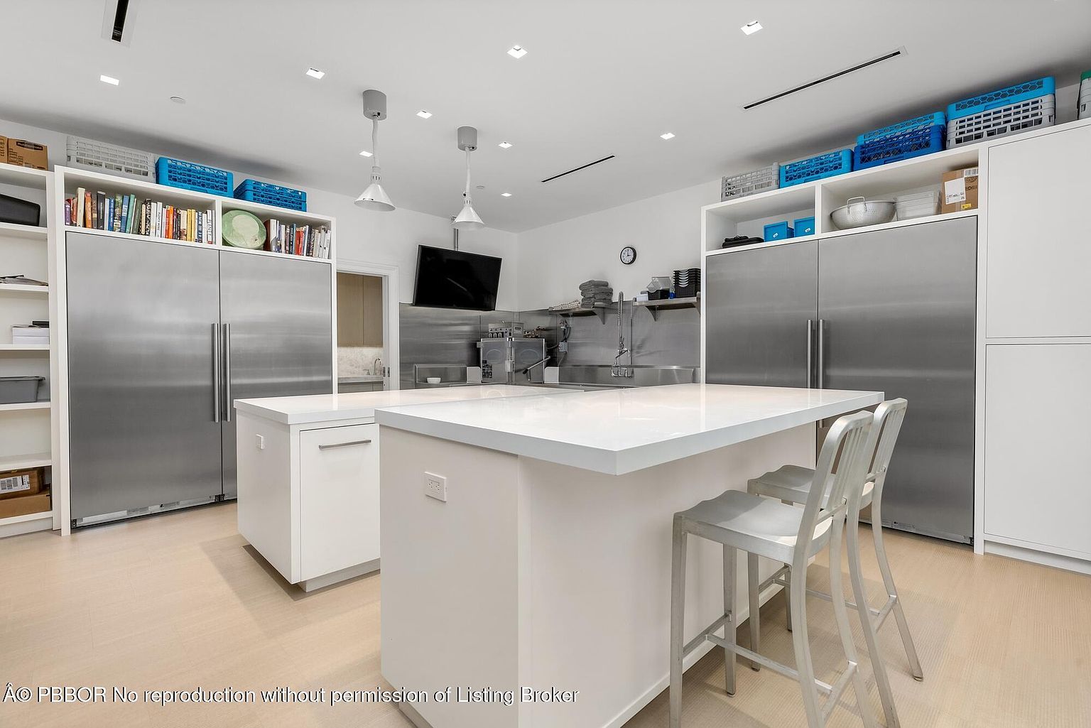 This is a bright and spacious kitchen featuring stainless steel appliances, including a large refrigerator, and white cabinetry. A large island with a white countertop provides ample workspace and seating with metal bar stools. The kitchen has a clean, modern aesthetic with recessed lighting and a stainless steel backsplash behind the sink area.