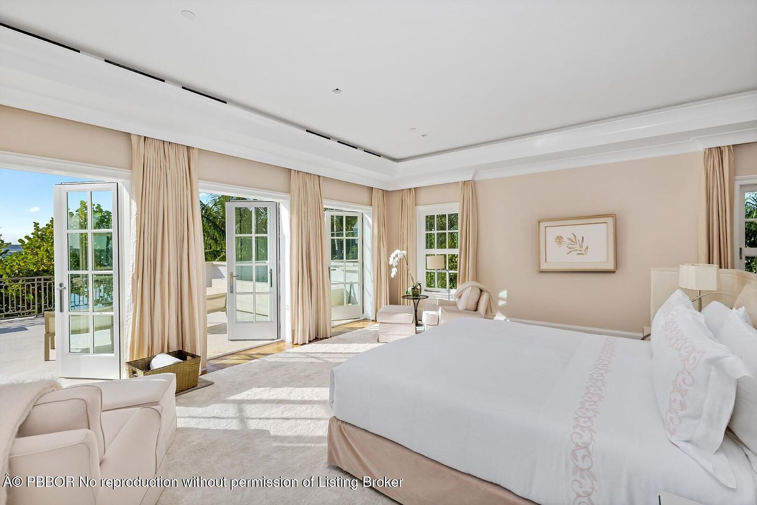 This is a bright and airy primary bedroom featuring a large bed with white linens and a light-colored upholstered headboard. The room has multiple sets of French doors leading to an outdoor balcony, complemented by neutral-toned curtains. The space is well-lit with natural light and features a soft, neutral color palette, creating a serene and luxurious atmosphere.