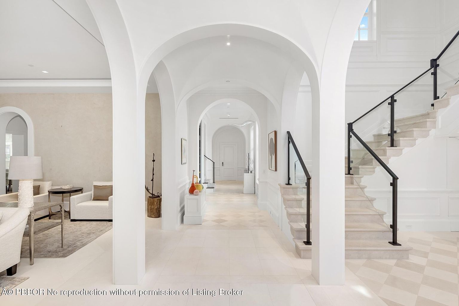 This interior shot showcases a grand hallway with a series of arched openings leading to other rooms, creating a sense of depth and spaciousness. A staircase with glass railings and dark metal accents adds a modern touch, while the light-colored tile flooring and white walls contribute to a bright and airy atmosphere. The design emphasizes clean lines and elegant simplicity, making it a sophisticated and inviting space.