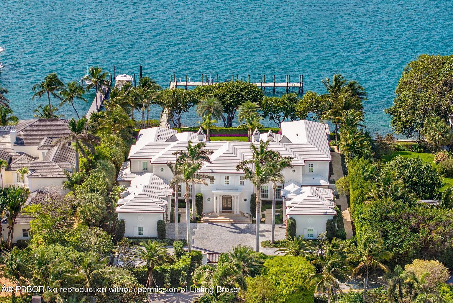 This aerial shot showcases a grand, white mansion nestled amidst lush greenery and bordering the ocean. The property features a meticulously maintained landscape with palm trees, a private dock extending into the water, and a large driveway leading to the entrance. The overall impression is one of luxury, privacy, and stunning waterfront living.