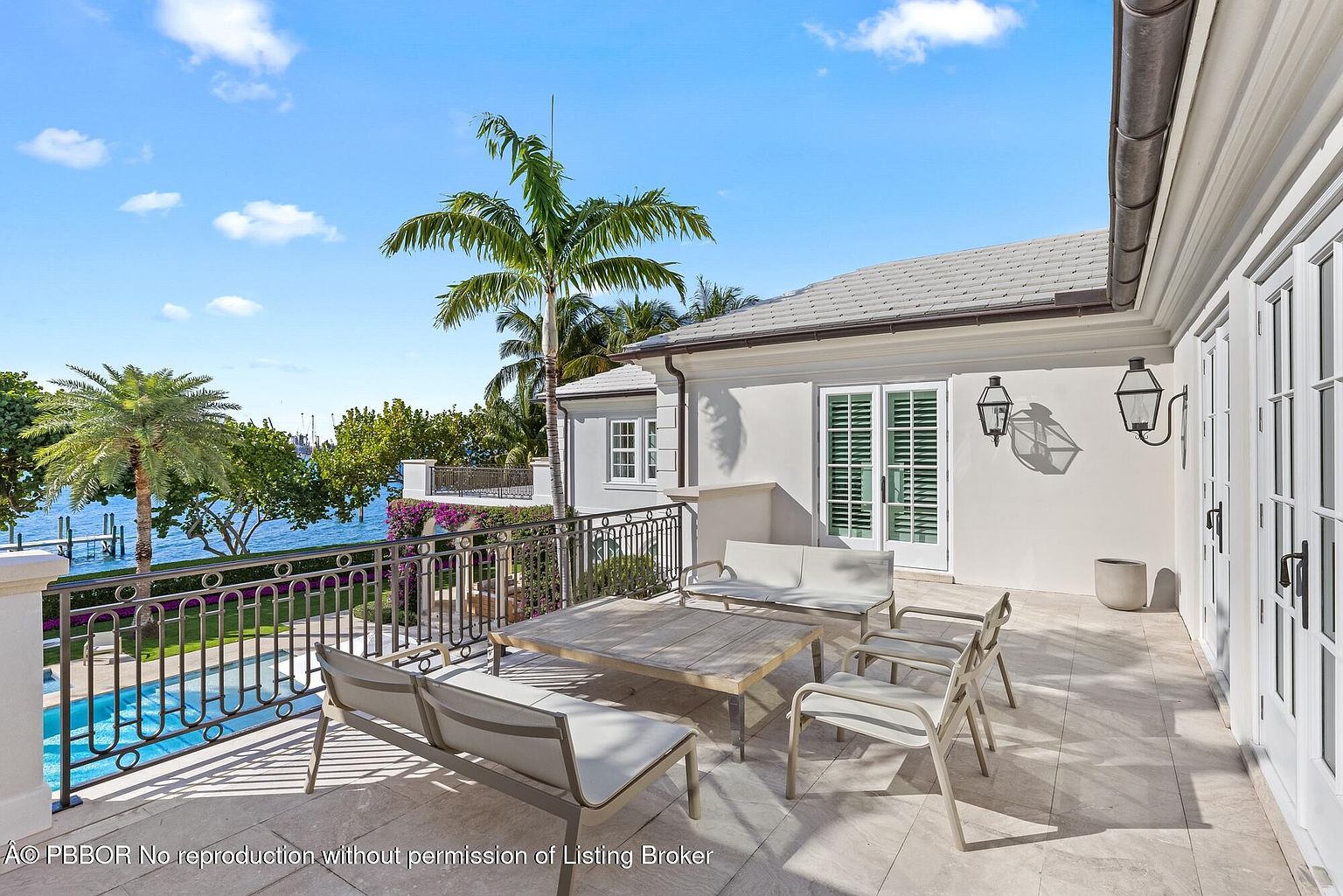 This image showcases a luxurious outdoor patio or balcony area, featuring stylish patio furniture including a sofa, chairs, and a coffee table. The space overlooks a beautiful waterfront view with palm trees and a swimming pool, creating a serene and upscale ambiance. The architecture is modern with white walls and dark accents, enhancing the overall elegance of the property.