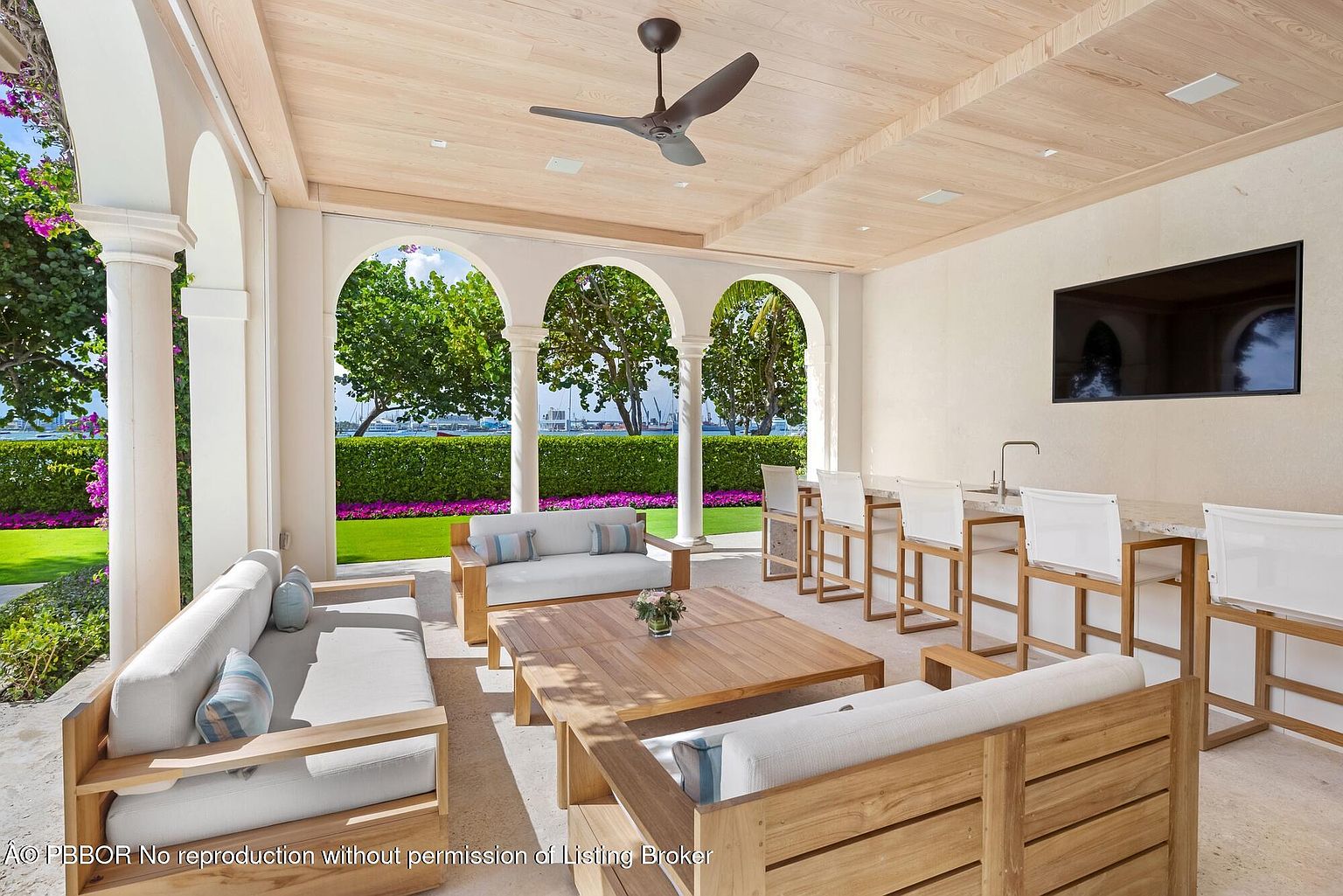 This image showcases a luxurious outdoor patio area, featuring comfortable seating arrangements with light-colored cushions and wooden frames. The space is partially covered with a light wood ceiling and supported by elegant white columns and arches, offering a view of a well-manicured lawn and waterfront. A bar area with high stools and a mounted television adds to the entertainment possibilities of this outdoor living space.