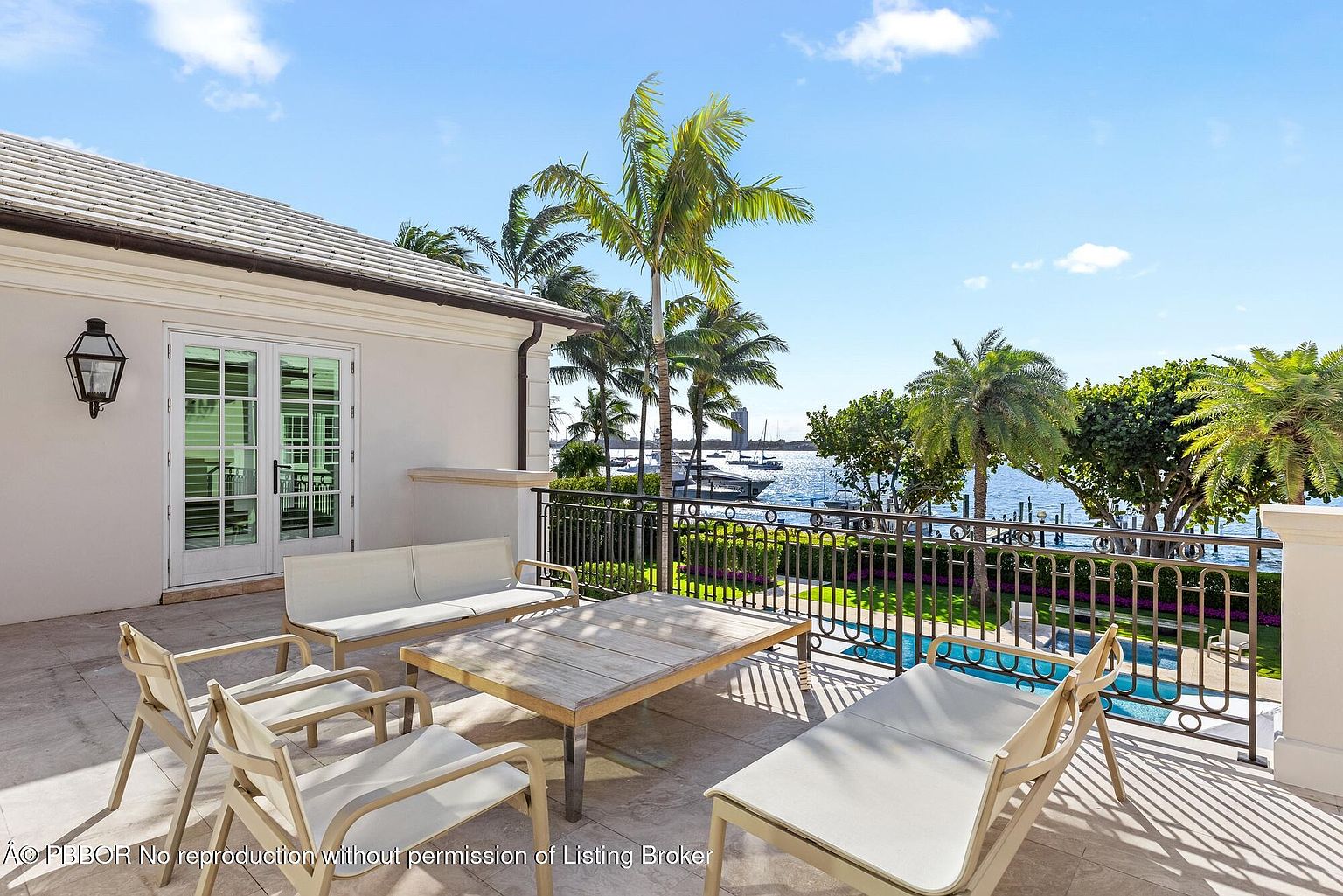 This image showcases a luxurious patio or balcony area with elegant outdoor furniture, including a sofa, chairs, and a coffee table. The space overlooks a serene waterfront view with palm trees and boats, suggesting a high-end coastal property. The architecture features a classic design with clean lines and a neutral color palette, enhancing the overall sophisticated ambiance.