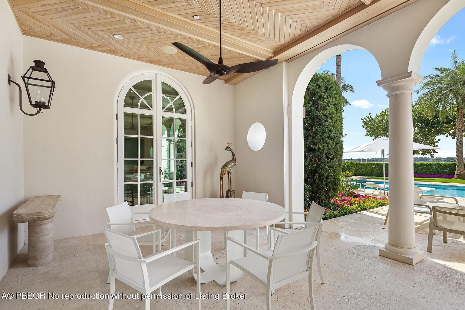 This image showcases a luxurious outdoor patio area, featuring a round dining table with white chairs, set against a backdrop of elegant architectural arches and a view of a swimming pool. The patio is well-lit and appears to be a perfect space for outdoor dining and relaxation, with a ceiling fan providing air circulation. The view through the arches reveals a well-maintained garden and pool area, enhancing the property's appeal.
