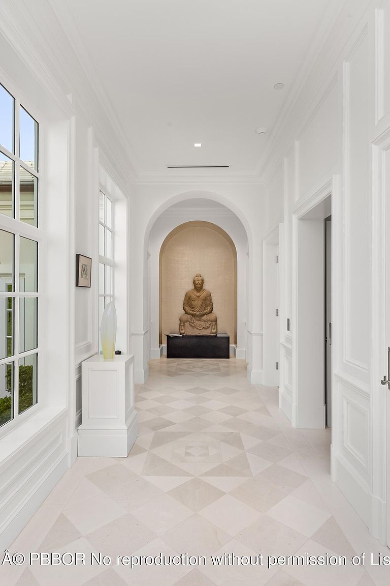 This is an interior shot of a bright, elegant hallway featuring a neutral color palette and a statue at the end of the hall. The hallway is lined with white walls, detailed molding, and arched doorways, leading to a focal point with a Buddha statue placed within an arched alcove. The flooring is a patterned tile, adding visual interest to the space.
