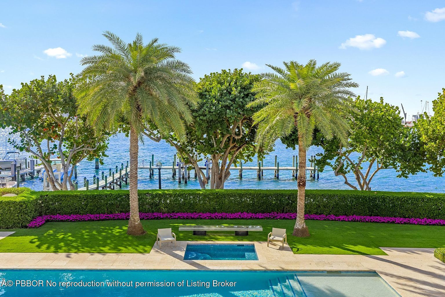 This image showcases a luxurious backyard with a pool and spa, framed by lush greenery and palm trees. A well-manicured lawn leads to a waterfront view, complete with a dock and boats in the distance. The scene exudes tranquility and upscale living, perfect for relaxation and entertainment.