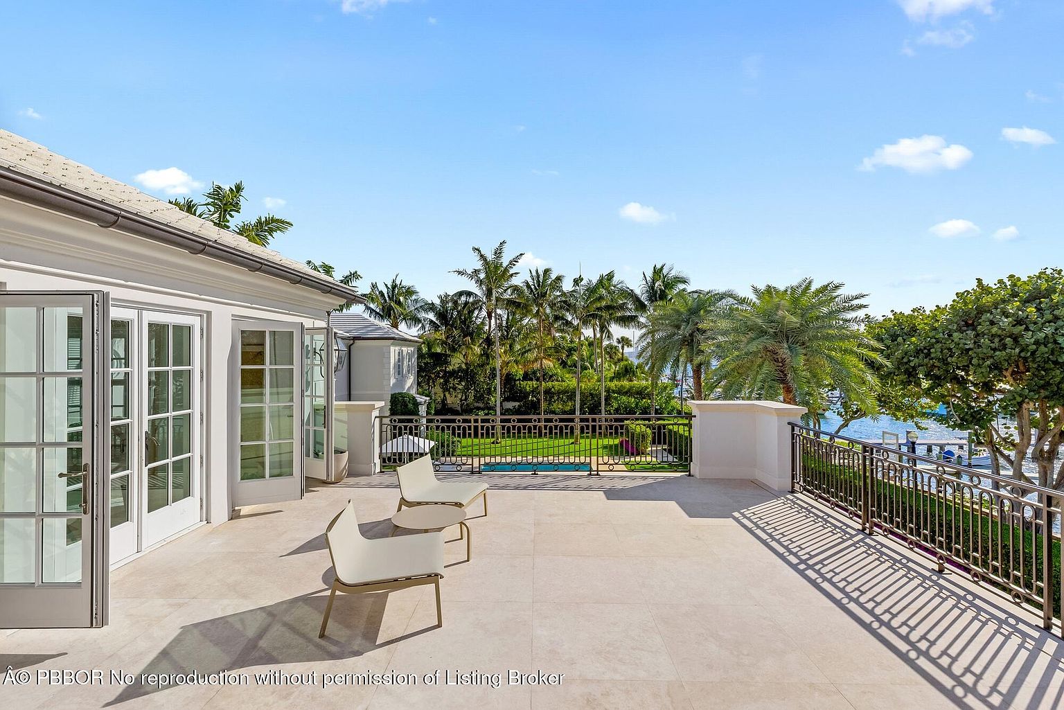 This image showcases a spacious patio or balcony area with two modern chairs, offering an inviting outdoor living space. The area features a tiled floor, white walls, and a decorative metal railing that overlooks a lush garden with palm trees and a glimpse of the water. The open French doors suggest easy access to the interior, enhancing the indoor-outdoor flow of the property.