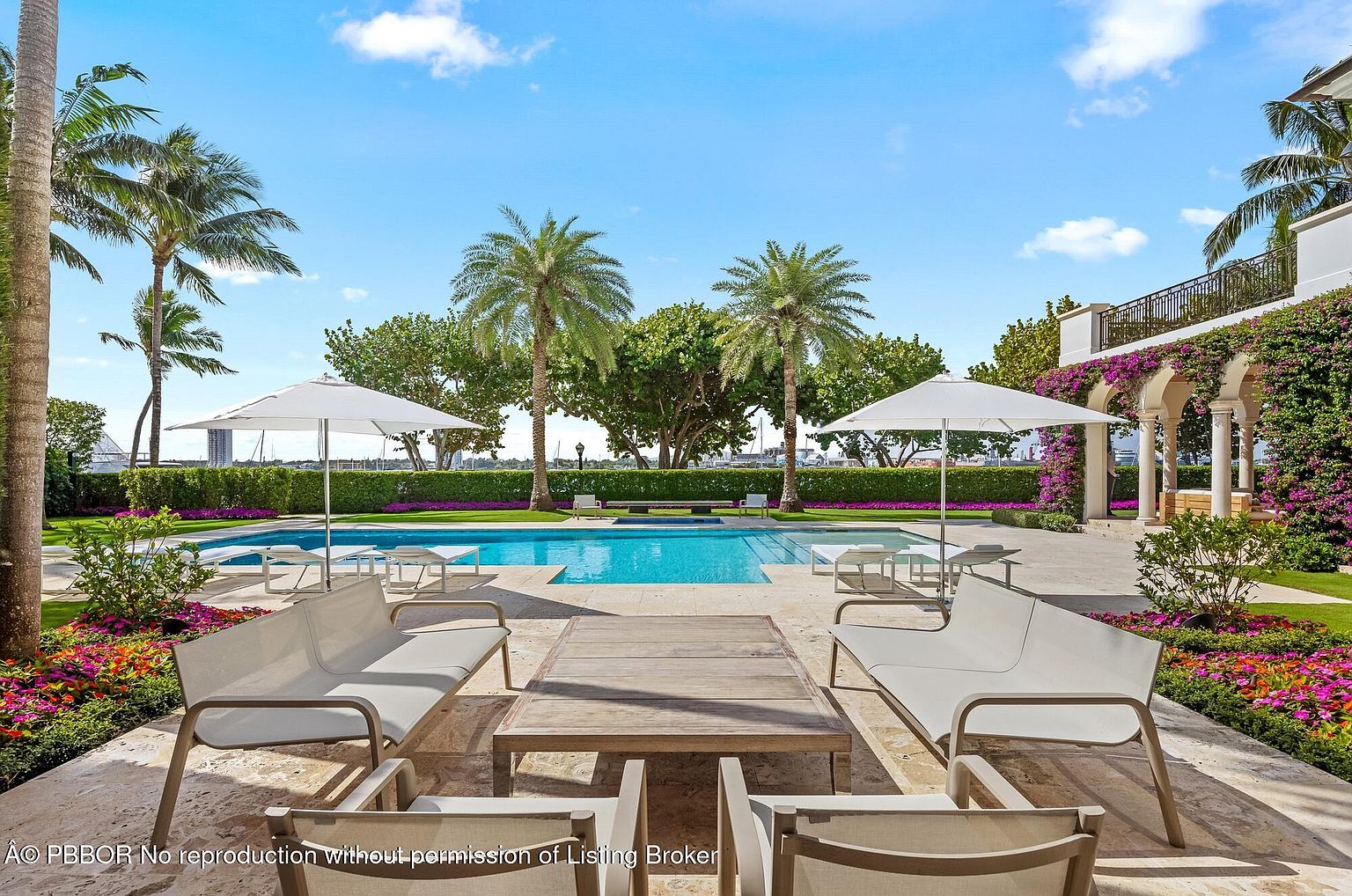 This image showcases a luxurious outdoor pool area, featuring a pristine pool surrounded by elegant patio furniture, including lounge chairs and a wooden table. The scene is framed by lush greenery, including palm trees and manicured hedges, with a glimpse of a building adorned with flowering vines in the background. The overall impression is one of tranquility and upscale outdoor living.