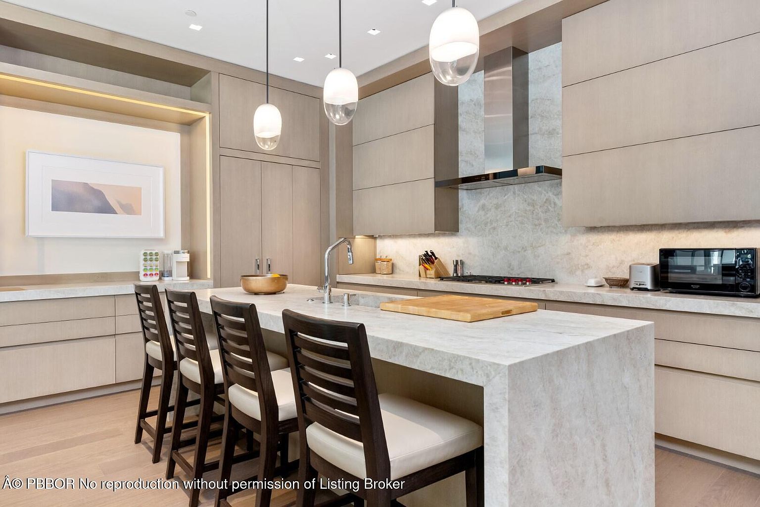 This is a well-lit, modern kitchen featuring light wood cabinetry and a large island with a light-colored countertop. The kitchen is equipped with stainless steel appliances and pendant lighting. The perspective is from a medium distance, showcasing the island and the surrounding cabinetry.