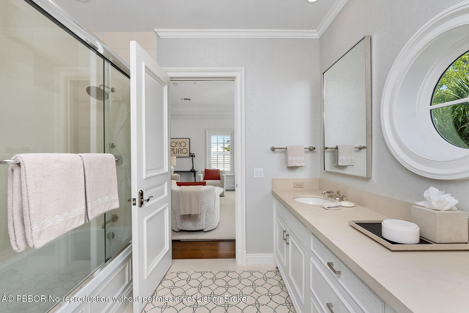 This is a well-lit primary bathroom featuring a glass-enclosed shower, a white vanity with a light countertop, and a round window. The walls are a light gray with a subtle pattern, and the floor has a geometric tile design. The open doorway leads to another room, creating a sense of spaciousness.
