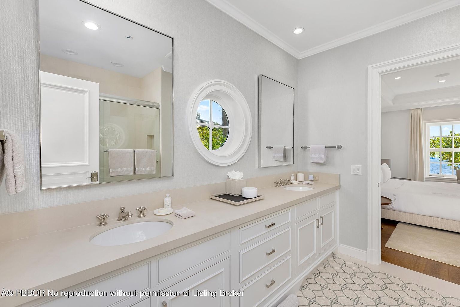 This is a well-lit primary bathroom featuring a double vanity with a light-colored countertop and white cabinetry. Two mirrors hang above the sinks, and a round window provides natural light. The bathroom opens into a bedroom, creating a seamless and luxurious transition between spaces.