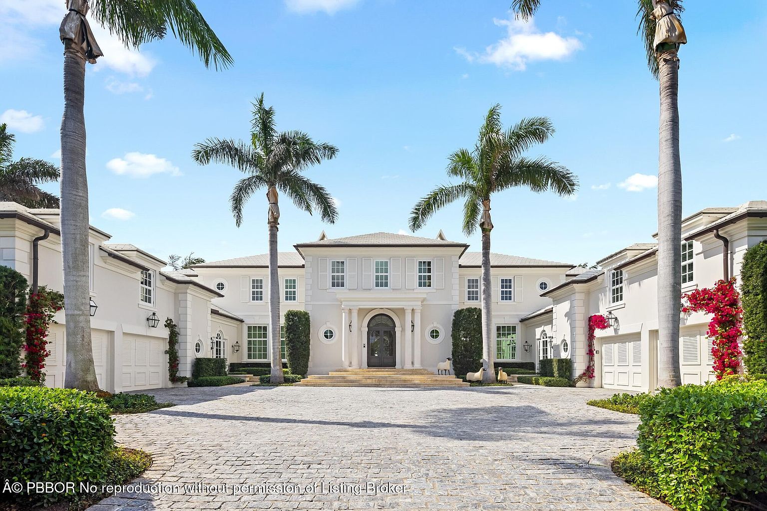 This is a front view of a grand, symmetrical two-story house with a light-colored facade, multiple windows with shutters, and a prominent central entrance framed by columns. The property features a circular driveway paved with stones, mature palm trees, manicured greenery, and attached garage wings on either side of the main structure. The overall impression is one of luxury and stately elegance.