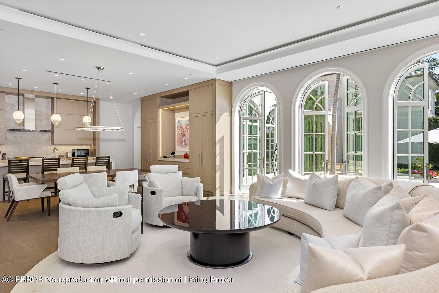 This is an interior shot of a luxurious living room that seamlessly blends with the kitchen and dining area. The living room features a curved sofa adorned with plush pillows, complemented by two armchairs and a sleek, dark-colored round coffee table. Large arched windows provide ample natural light and a view of the outdoors, enhancing the room's airy and sophisticated ambiance.