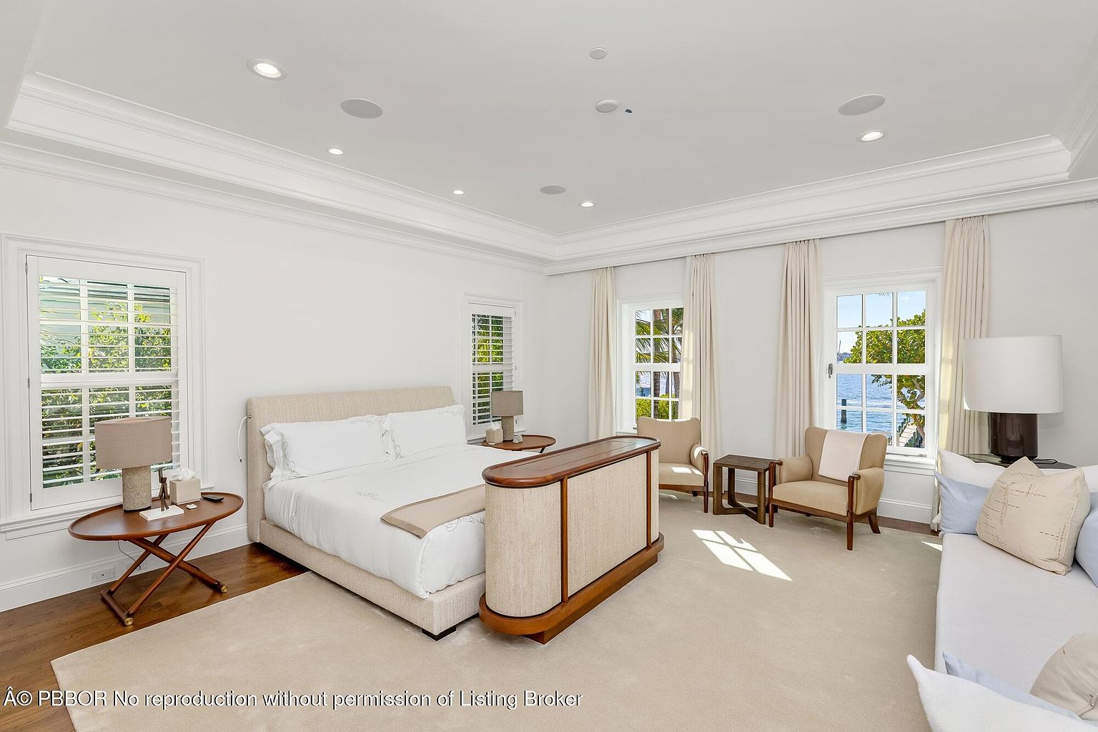 This is a bright and spacious primary bedroom featuring a large bed with neutral linens, flanked by side tables and lamps. The room is well-lit with natural light streaming through the windows, complemented by recessed lighting. The decor is minimalist and elegant, creating a serene and inviting atmosphere.