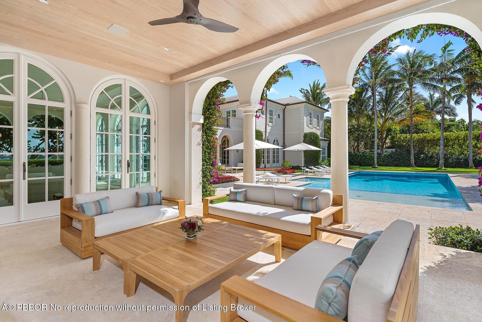 This image showcases a luxurious outdoor patio area with elegant architectural details. The patio features comfortable seating arrangements with plush cushions, a wooden coffee table, and arched openings that frame a view of the pool and lush landscaping. The overall impression is one of sophisticated outdoor living and relaxation.