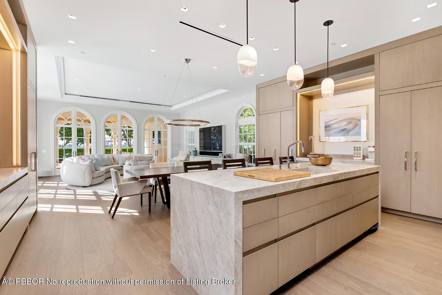 This interior shot showcases a modern, open-concept kitchen and dining area. The kitchen features a large island with a marble countertop and light wood cabinetry, complemented by pendant lighting. The dining area includes a wooden table and chairs, with arched windows providing natural light and a view of the outdoors, creating a bright and inviting space.