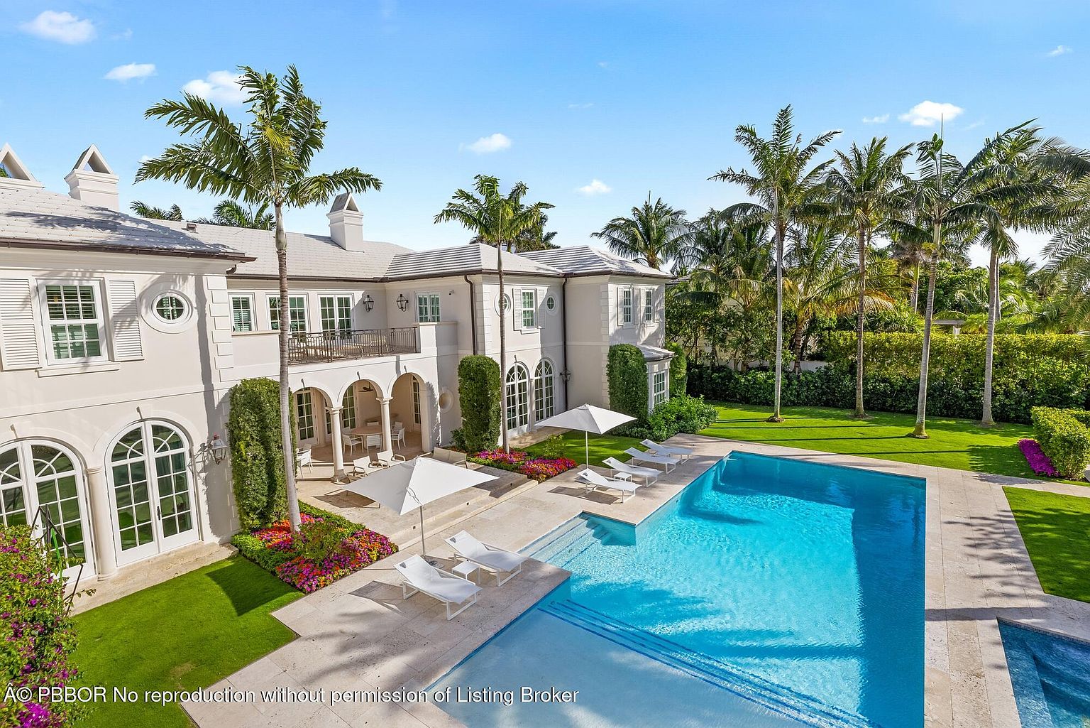 This aerial view showcases a luxurious estate with a pristine pool, lush green lawns, and meticulously landscaped gardens. The elegant architecture of the home is complemented by tall palm trees, creating a serene and upscale ambiance. The property exudes sophistication and offers a private oasis for relaxation and entertainment.